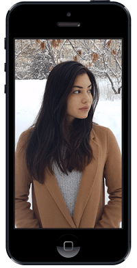 Donna con cappotto marrone e capelli lunghi neri in un ambiente innevato, vista attraverso lo schermo di uno smartphone.